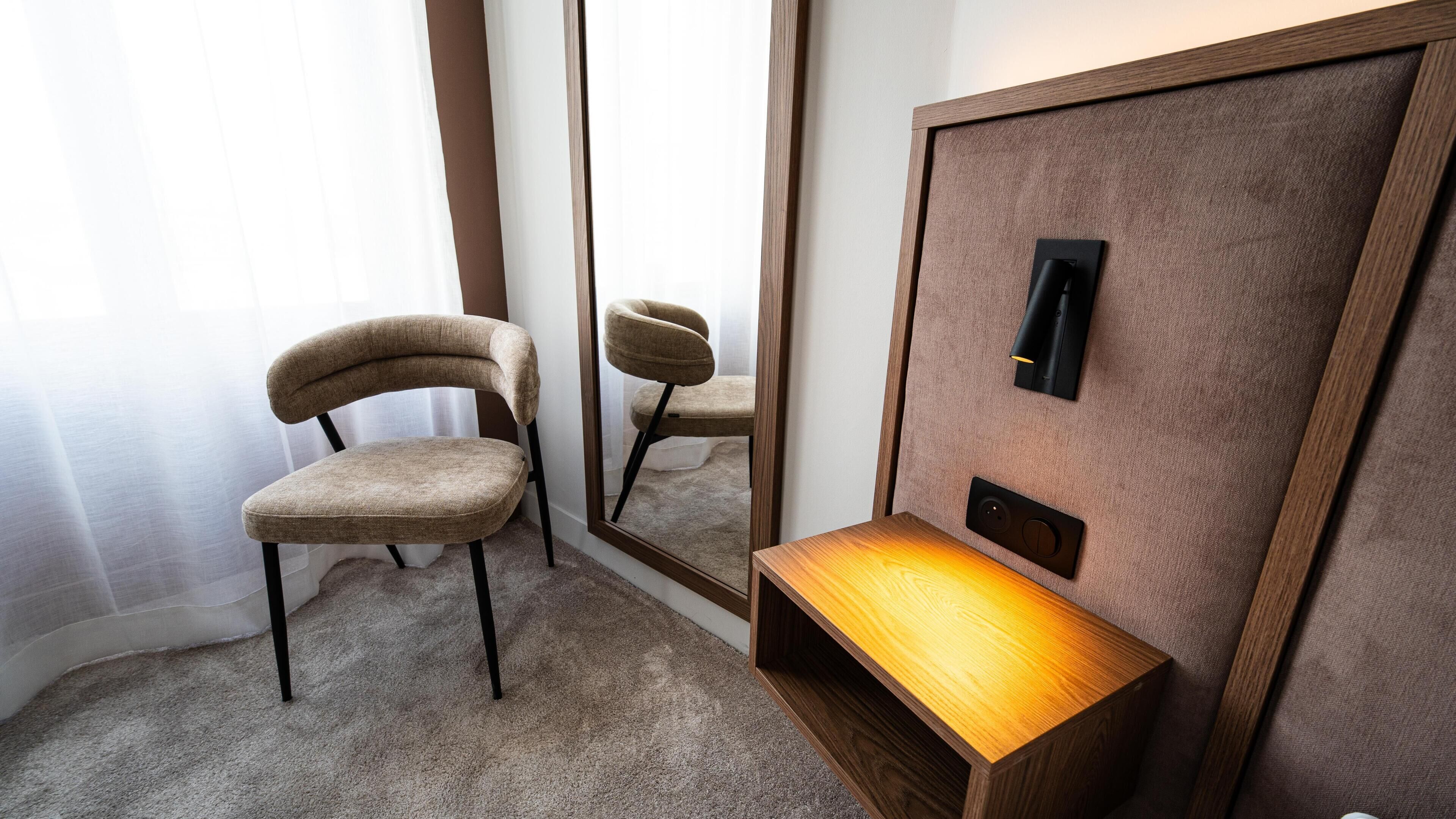 A room with a chair, a mirror, a bedside table, and a wall-mounted light fixture.