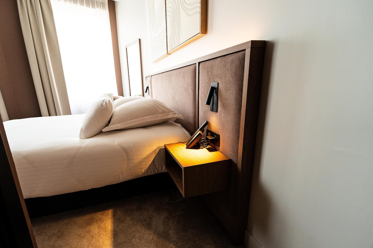 Una cama bien hecha con almohadas, una mesita de noche con lámpara, y un aplique de pared.