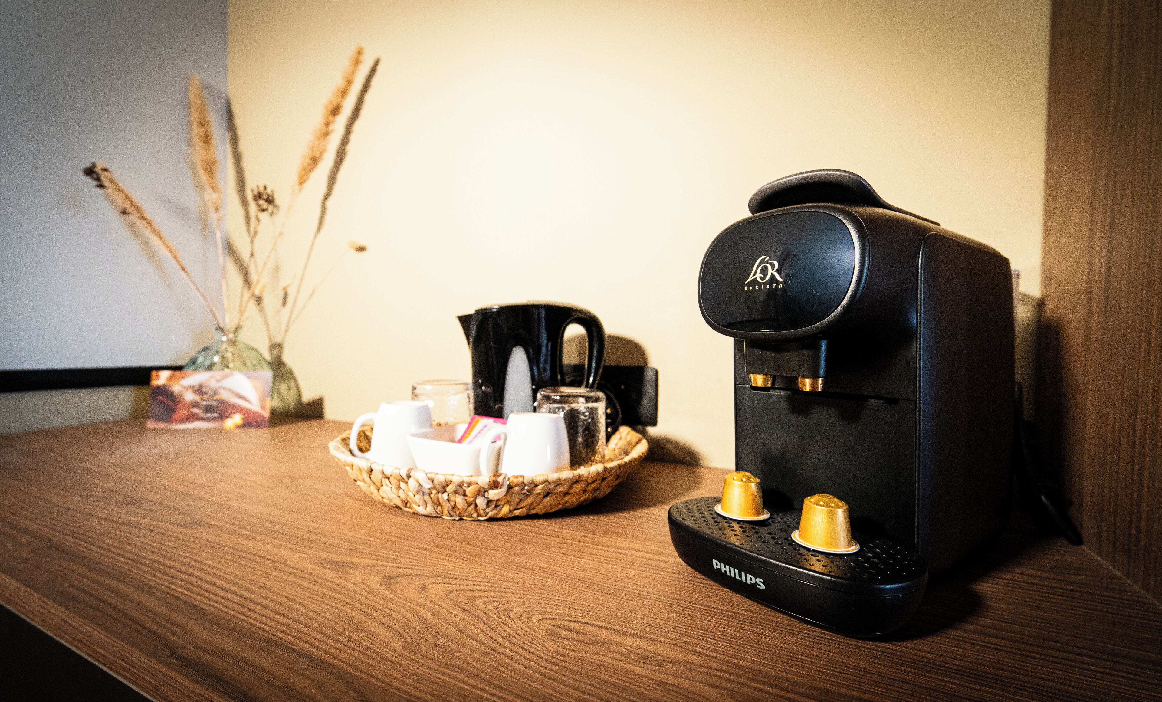 A coffee maker on a wooden surface with a basket of coffee cups and a vase of dried plants.