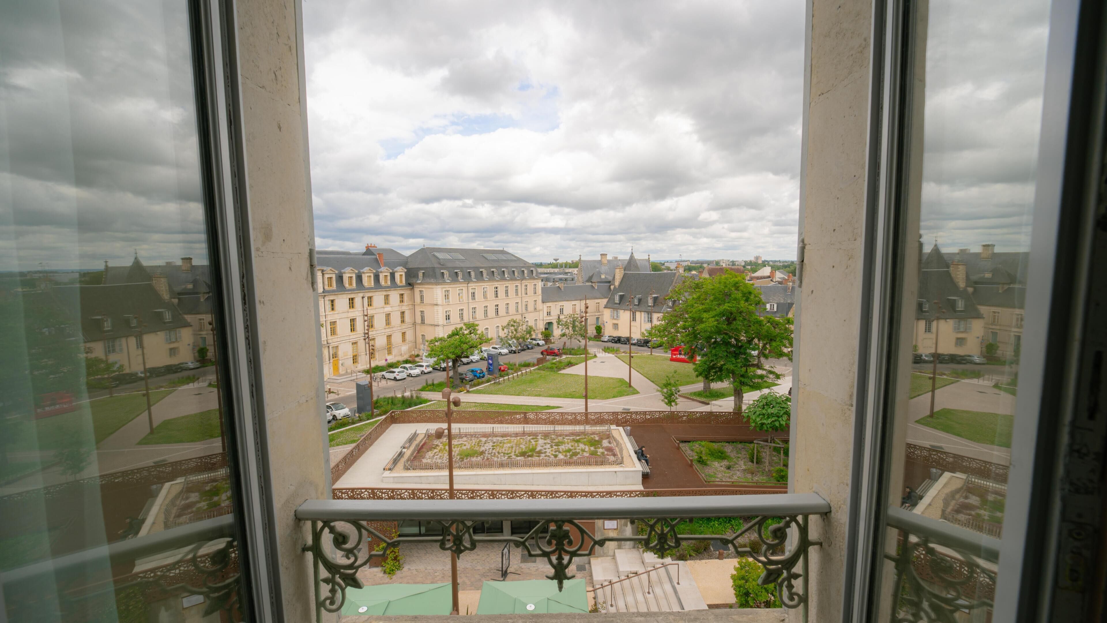 Une vue depuis une fenêtre donnant sur un balcon, avec vue sur une cour intérieure où se trouvent des bâtiments, un arbre et un véhicule rouge.