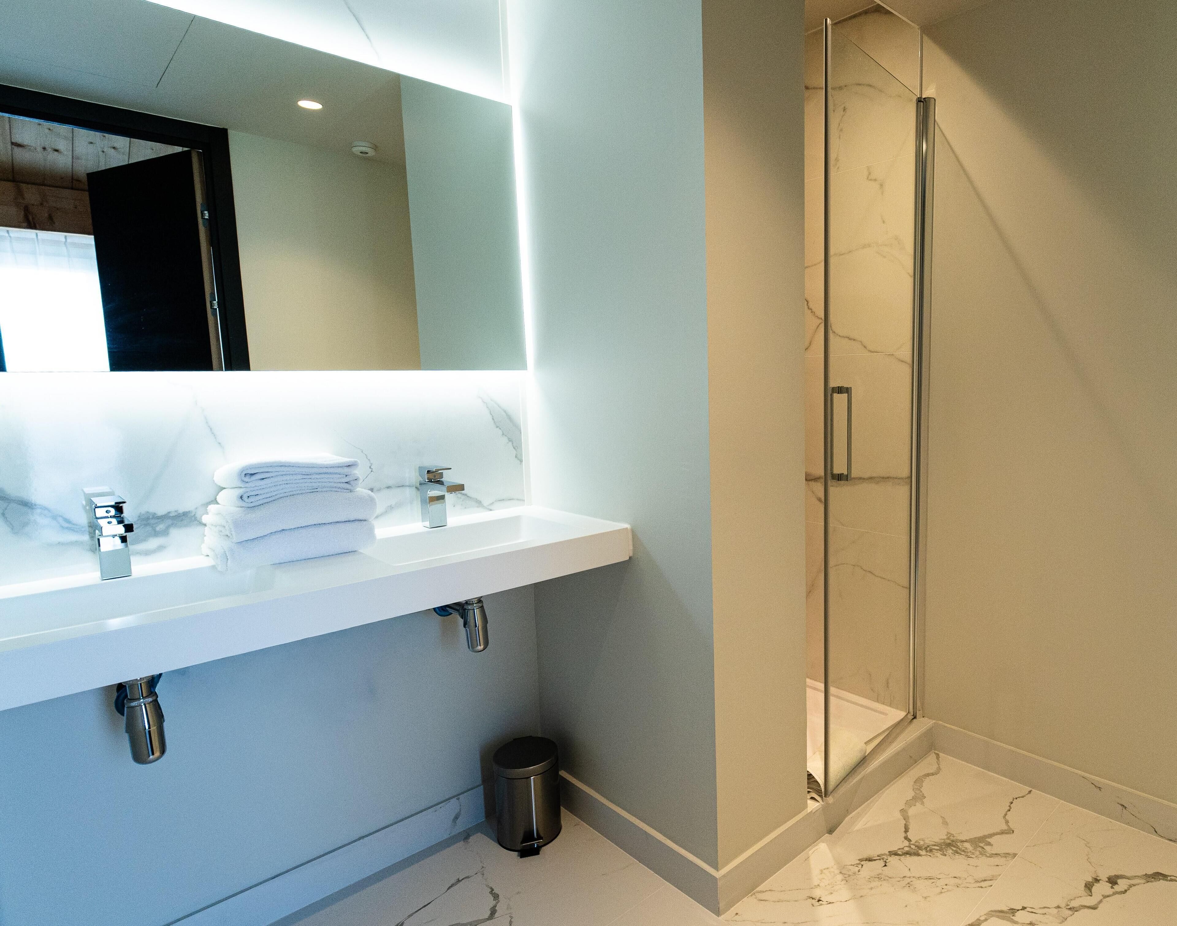 A modern bathroom with a white marble sink, a glass shower enclosure, and a visible trash bin.