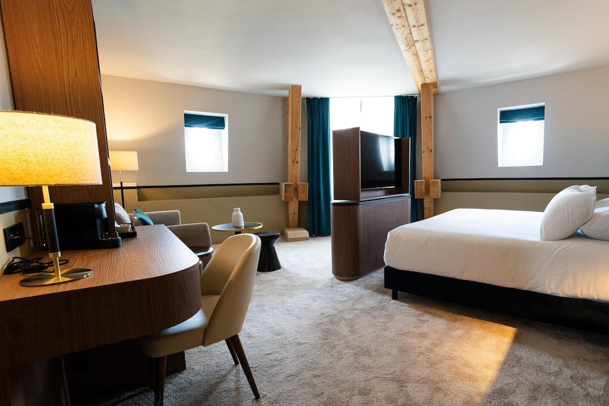 Habitación de hotel con cama, escritorio, silla, televisor y dos ventanas.