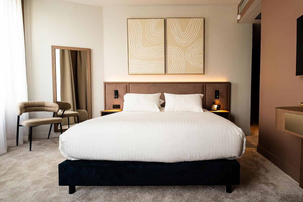 Habitación de hotel con una cama grande, dos mesitas de noche, una silla, un televisor de pantalla plana y arte abstracto en la pared.