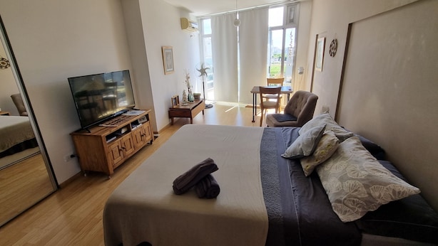 Un dormitorio con cama, televisor, mueble de madera, silla, escritorio y ventana con vistas.