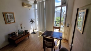 Una habitación con una mesa de madera, sillas, un ventilador de pie, un cuadro enmarcado y una ventana con vista a la vegetación.