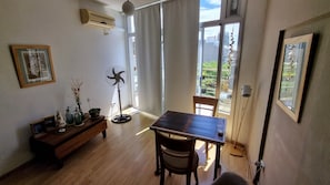 Una habitación con una mesa de madera, sillas, un ventilador de pie, un cuadro enmarcado y una ventana con vista a la vegetación.