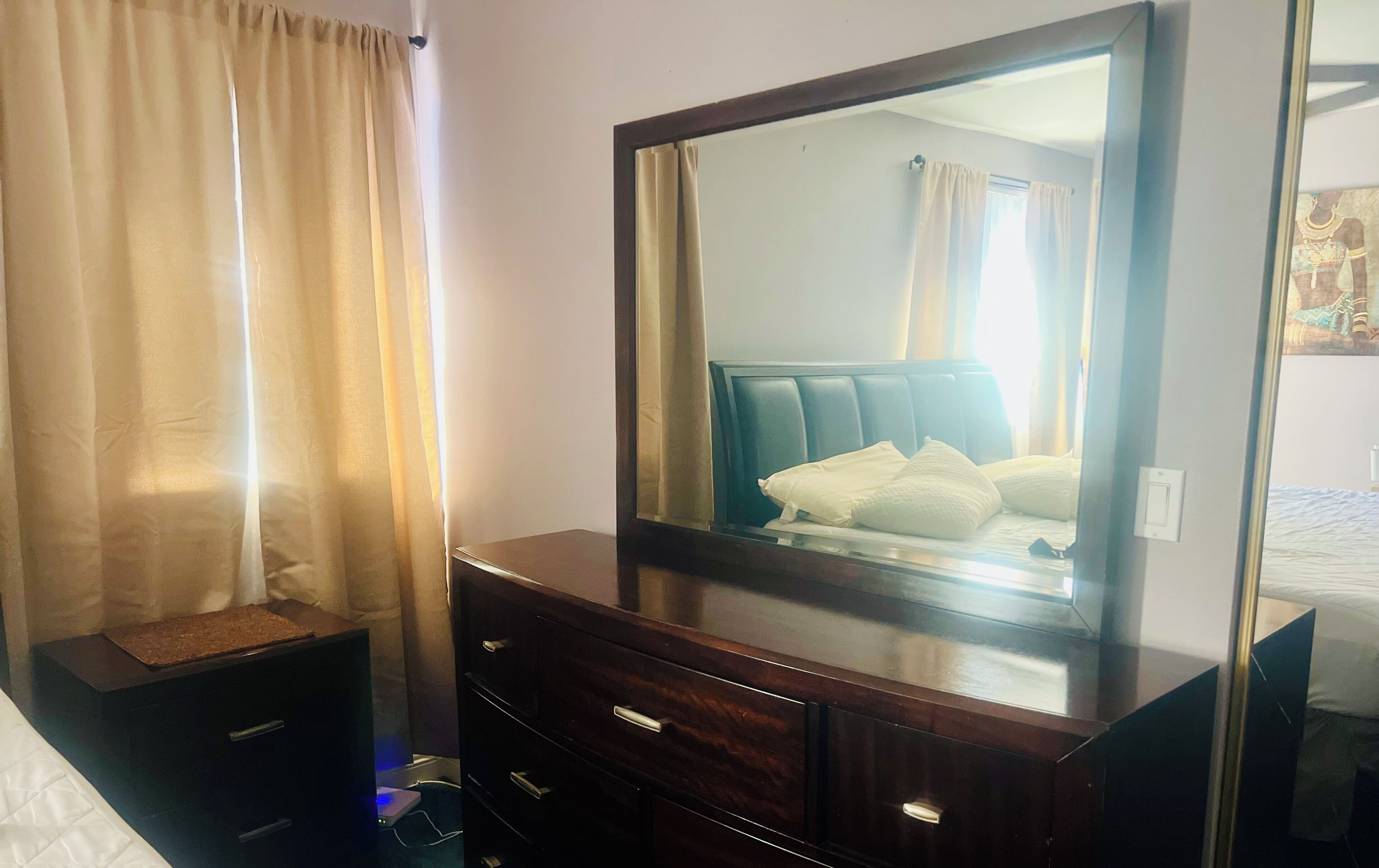 Una habitación de hotel con un espejo grande, una cama con ropa de cama blanca, un aparador de madera con cajones y una ventana con cortinas.