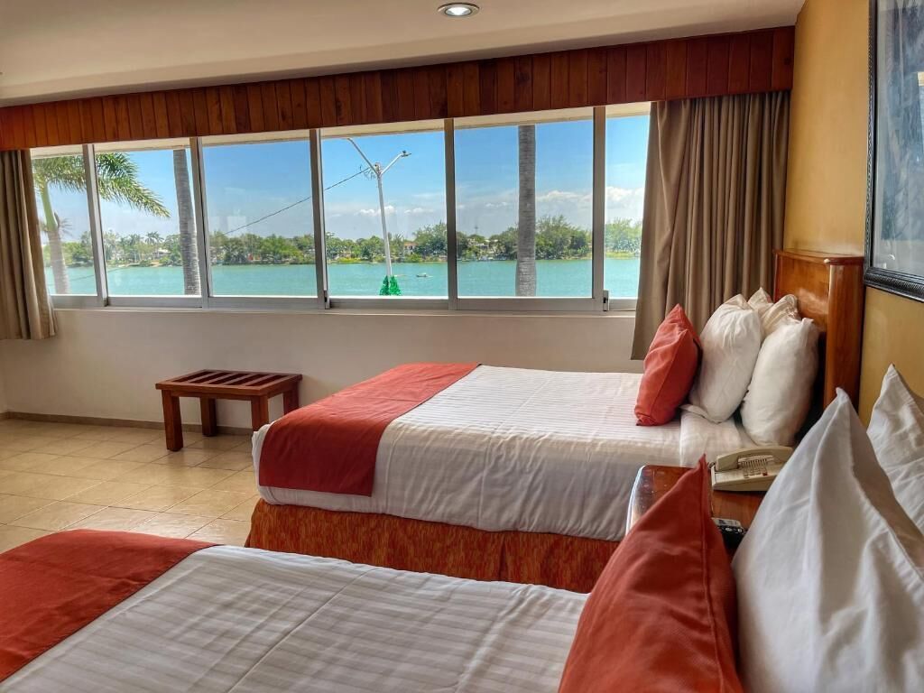 A hotel room with two beds, a view of a body of water, a small table, and a window with curtains.