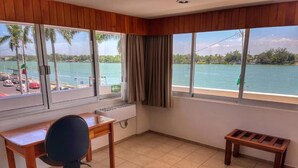 Una habitación con escritorio, silla y banco, ventanas amplias con vistas a un cuerpo de agua y palmeras.