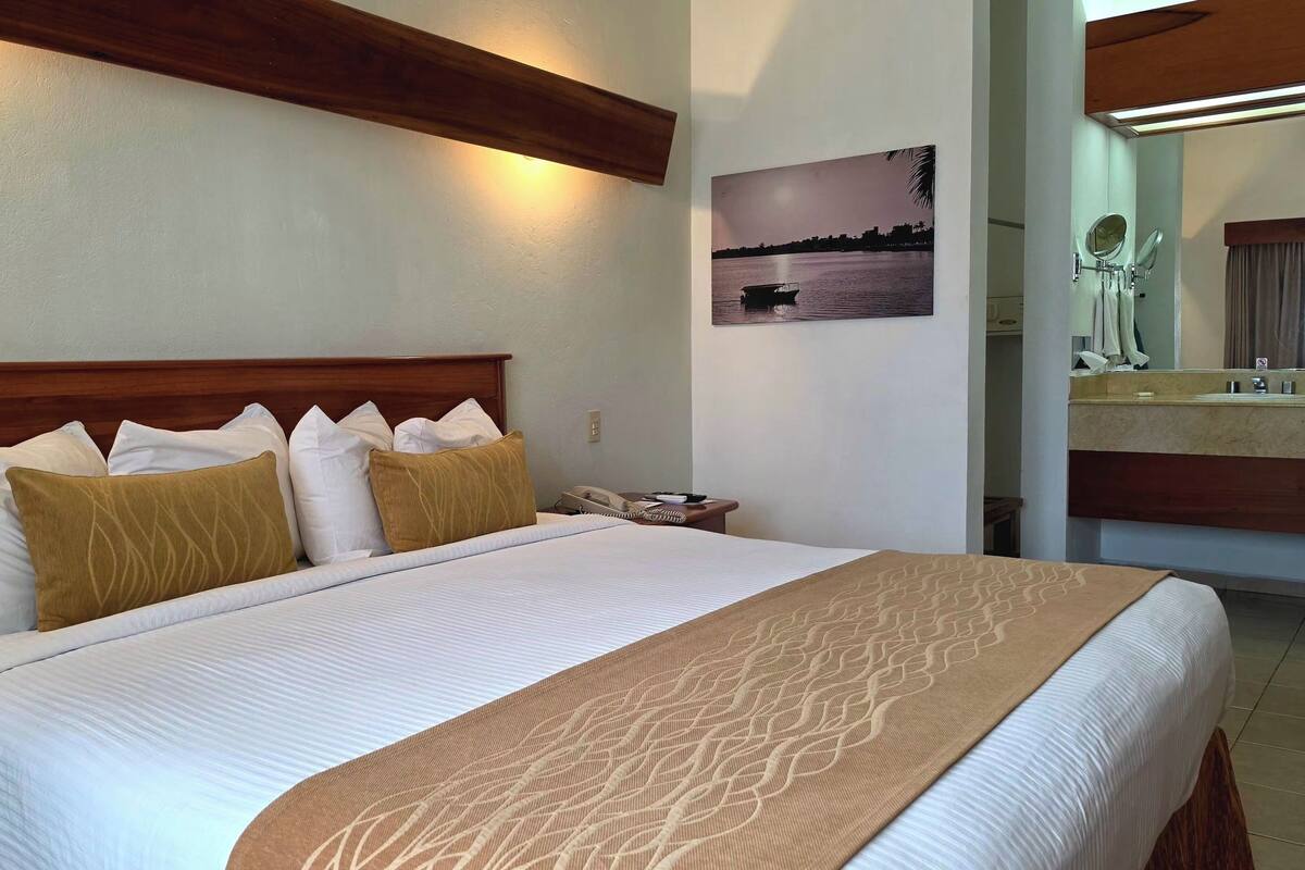 Habitación de hotel con cabecera de madera, cama con ropa de cama blanca y beige, teléfono junto a la cama, cuadro colgado en la pared y baño con espejo y perchero.