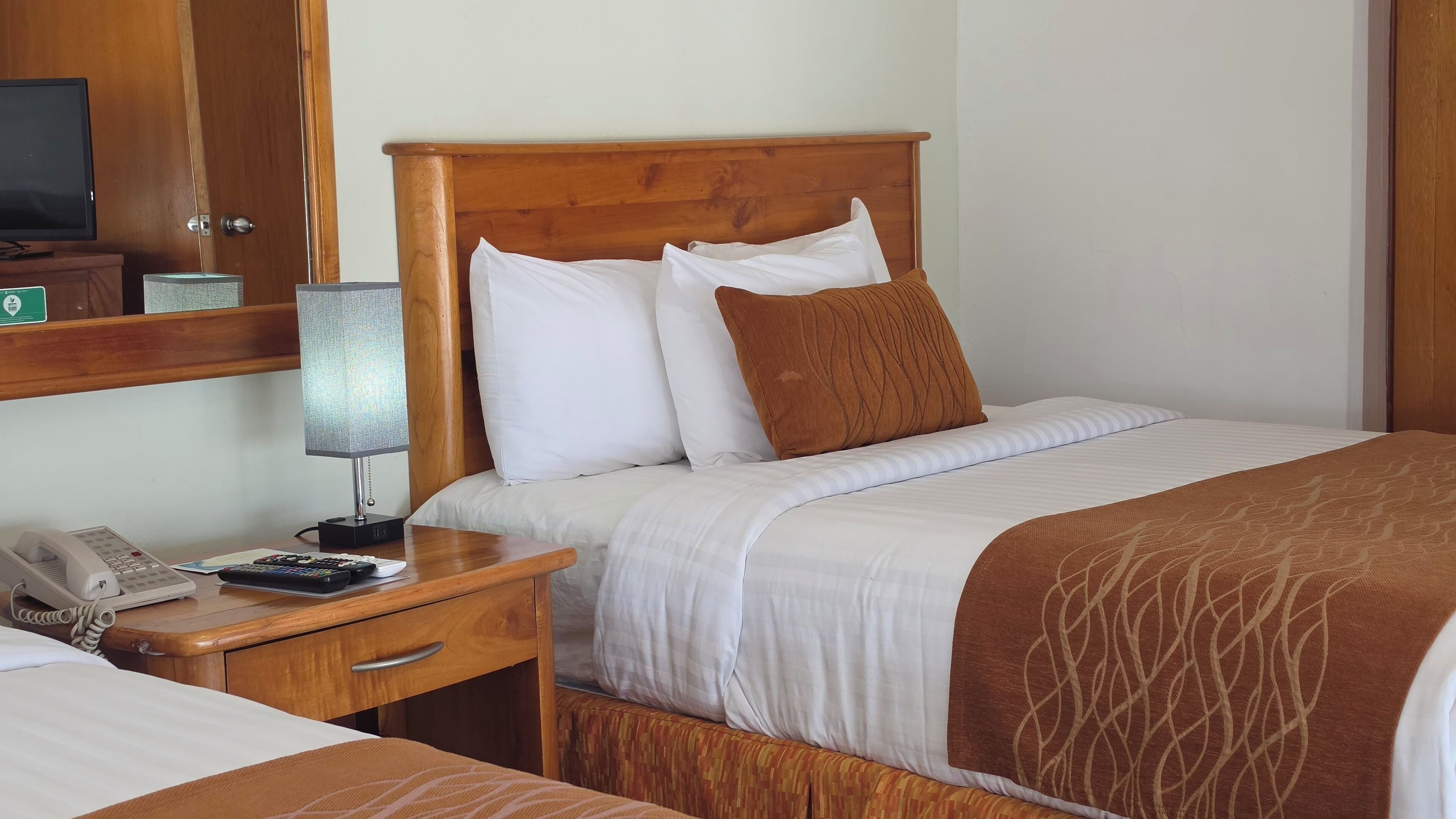 A hotel room with a wooden headboard, a bedside table with a lamp, a phone, and a bed with white and brown bedding.