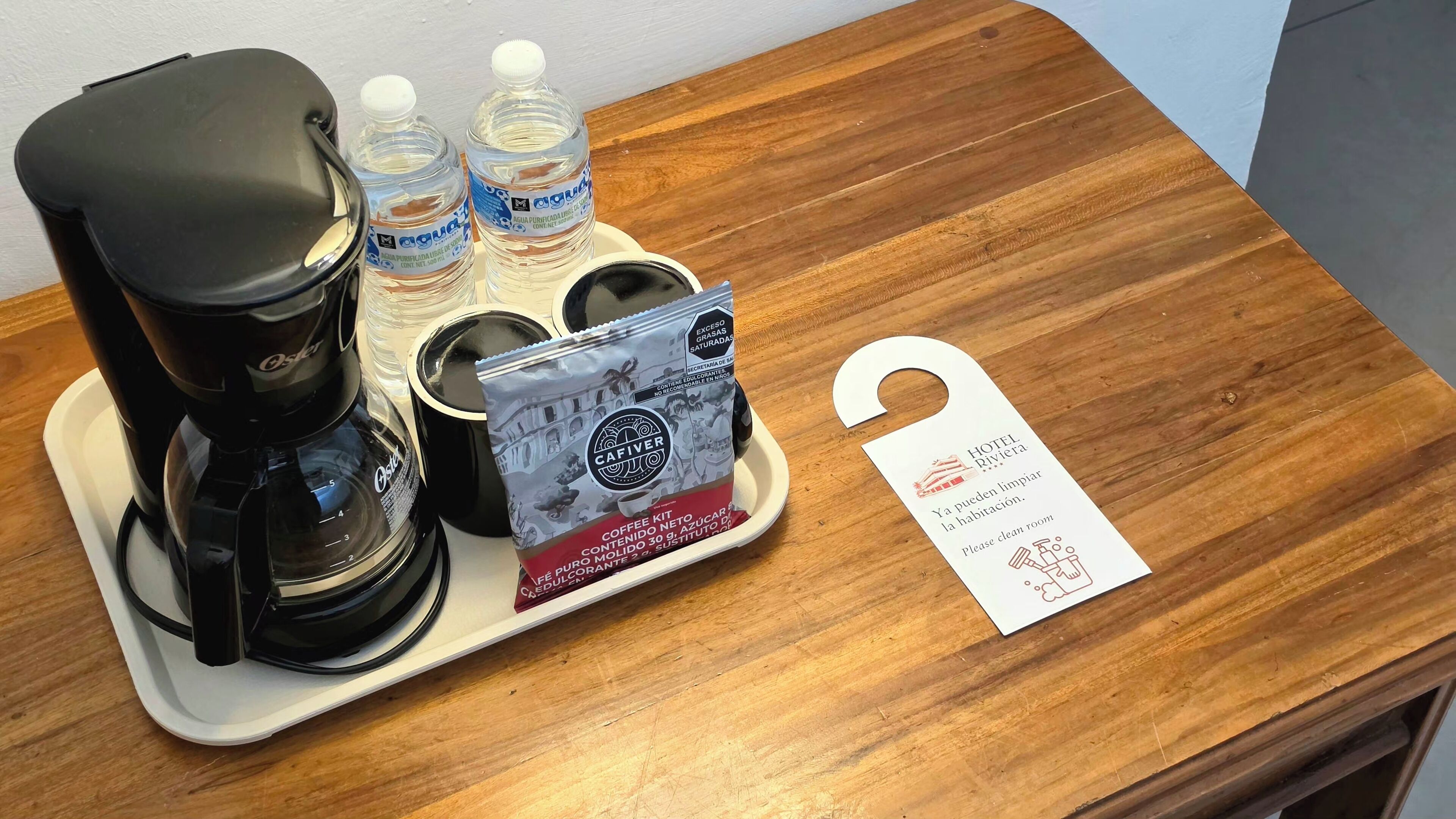 Una cafetera, dos botellas de agua, una taza de café, un paquete de café y un cartel con instrucciones de limpieza del cuarto sobre una mesa de madera.