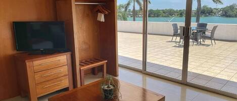 A room with a wooden desk, a television, a wardrobe, and a balcony with a table and chairs.