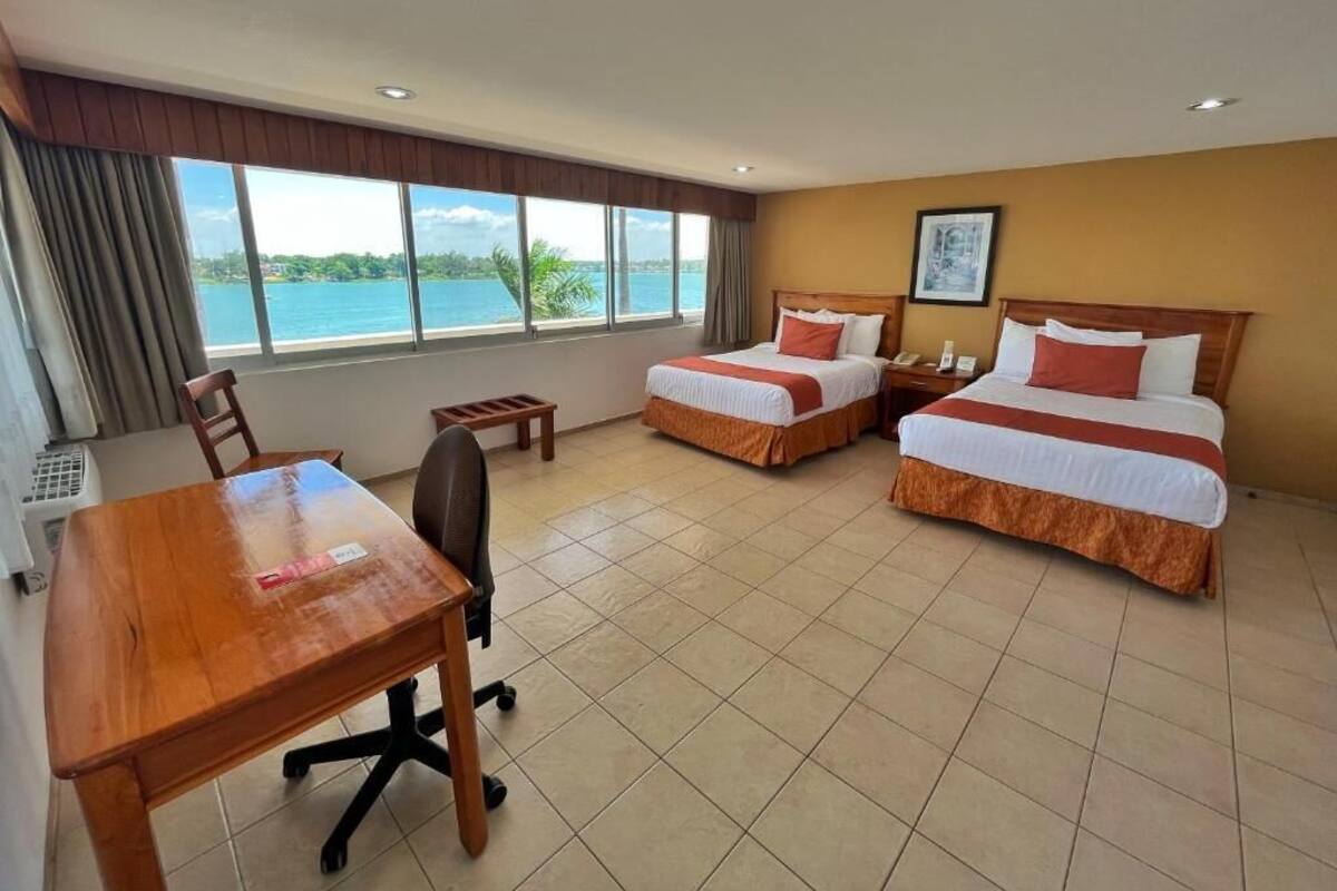 Habitación de hotel con dos camas, un escritorio de madera, una silla y un gran ventanal con vista al mar.