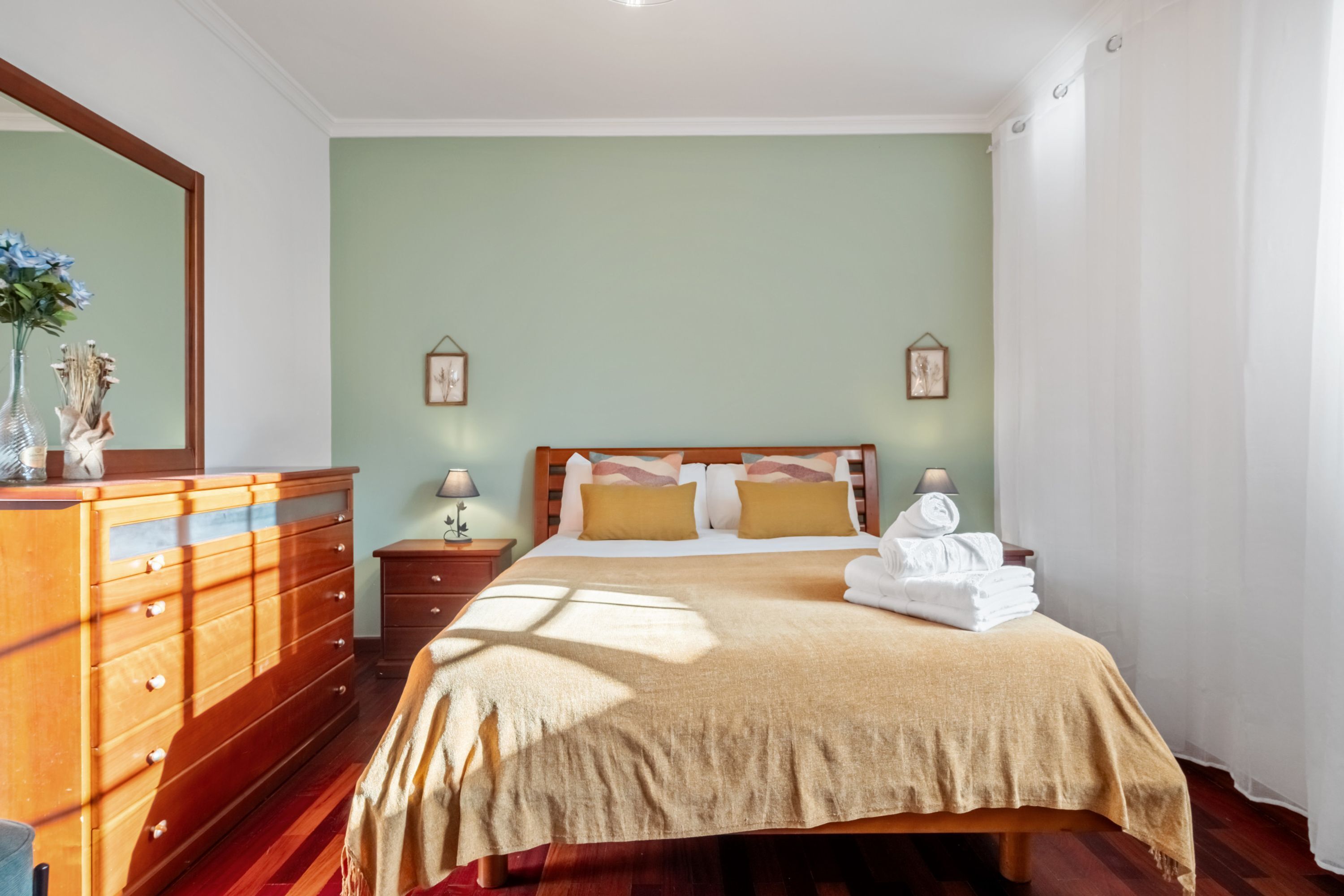 Un dormitorio con un aparador de madera, una cama con una manta color beige, dos lámparas de noche y un espejo en la pared.