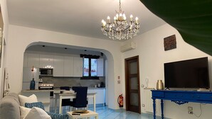 Ein Wohnzimmer mit einer Couch, eine Essecke mit Tisch und Stühlen, eine Küche mit Schränken und Geräten, ein Fernseher und eine Pendelleuchte.