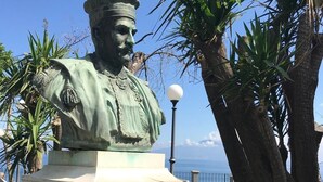 Eine Statue einer historischen Persönlichkeit mit Podestinschrift, umgeben von Grünpflanzen und blauem Himmel.