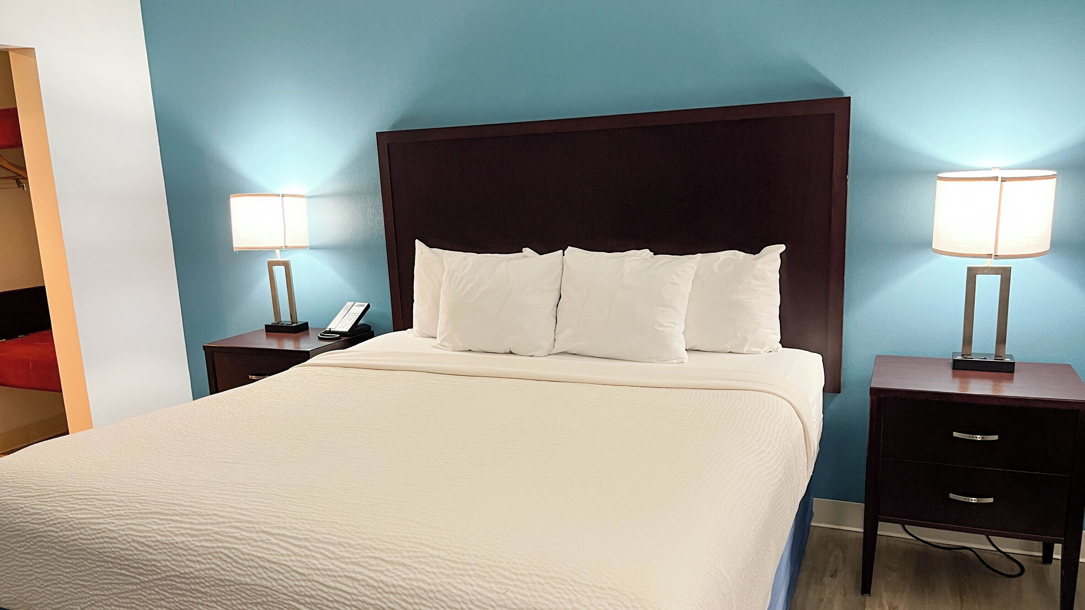 A hotel room with a large bed, two bedside tables with lamps, a dark headboard, and a blue wall.