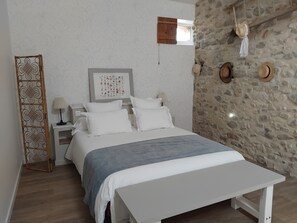 Een slaapkamer met een bed, nachtkastje, lamp, fotolijst, raam en een stenen muur.
