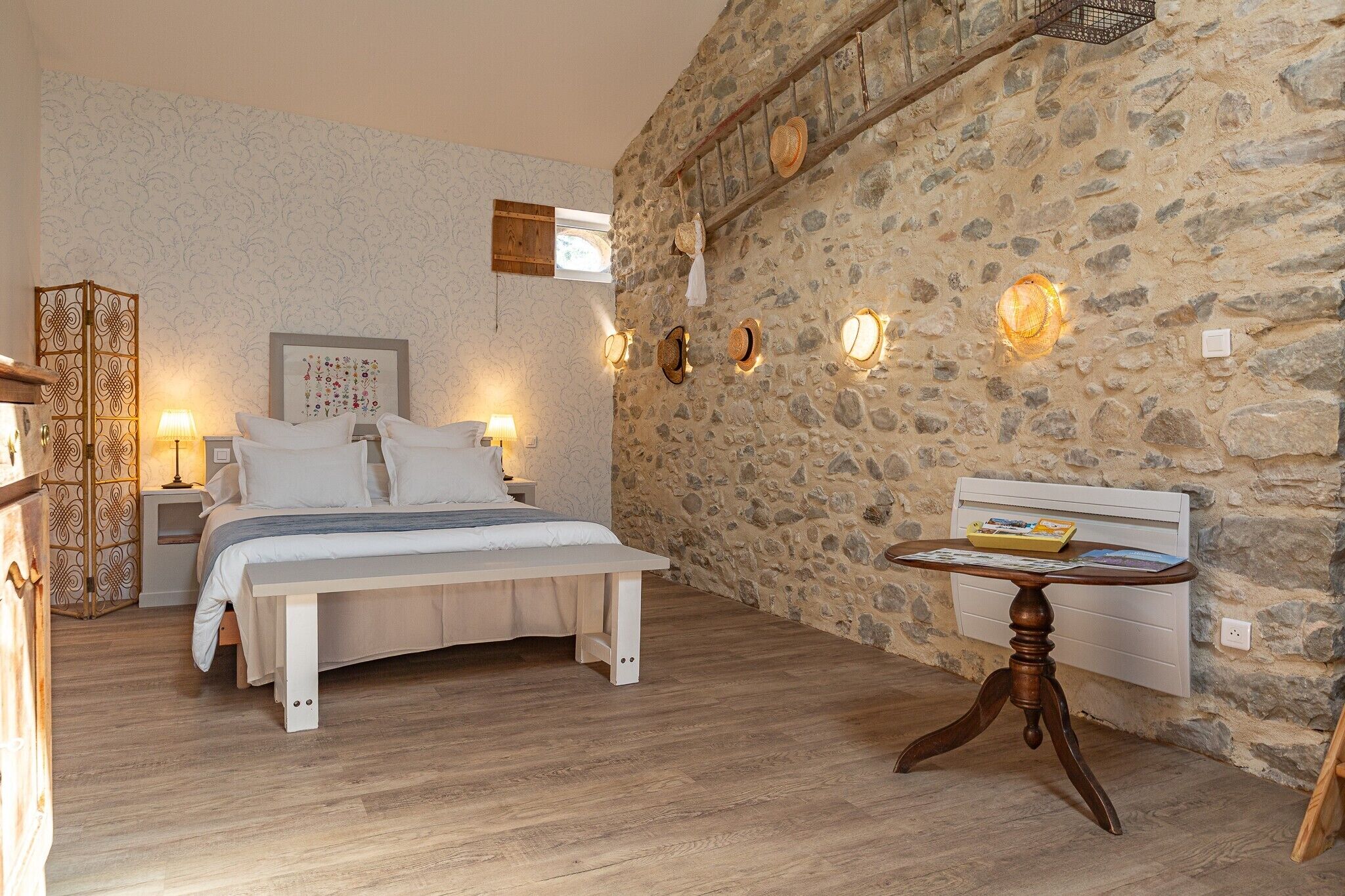 Een slaapkamer met een stenen muur, een bed met witte lakens, bedlampen en een klein tafeltje met een boek.