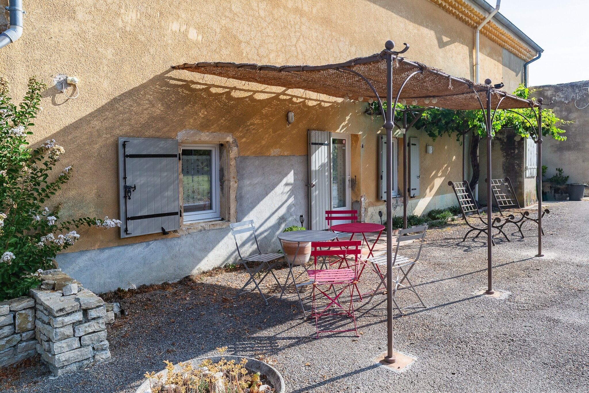 Un edificio con pared de piedra, una mesa y sillas rojas, y una pérgola de metal.