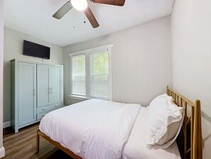 Ein Schlafzimmer mit einem Bett, einem Deckenventilator, einem an der Wand befestigten Fernseher und einem Fenster mit Jalousien.
