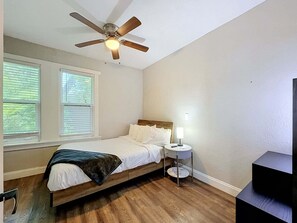 Ein Schlafzimmer mit einem Bett, einem Deckenventilator, einem Nachttisch und einem Fenster mit Jalousien.