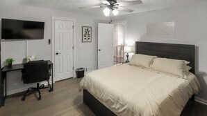 Ein Schlafzimmer mit Bett, Schreibtisch, Stuhl, Fernseher, Deckenventilator und einer Tür zu einem weiteren Raum.