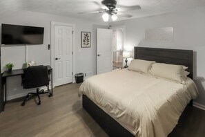 Un dormitorio con cama, escritorio, silla, televisor, ventilador de techo y una puerta que da a otra habitación.