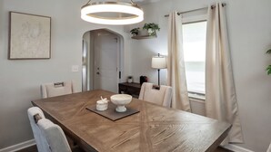 Ein Essbereich mit Holztisch, Stühlen, einer hängenden Leuchte, einem Wandbild und einem Fenster mit Vorhängen.