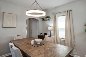 Un comedor con mesa de madera, sillas, lámpara colgante, un cuadro en la pared y una ventana con cortinas.