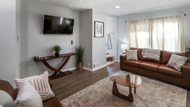 Ein Wohnzimmer mit braunem Ledersofa, einem Couchtisch, einem an der Wand montierten Fernseher und einem großen Fenster mit Vorhängen.