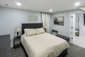 Ein Schlafzimmer mit einem Bett, Nachttischen, einer Bank, einem Gemälde und einer Tür zu einem Badezimmer.