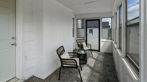 Ein Korridor mit einer weißen Tür, schwarzen Korbstühlen, einem kleinen runden Tisch mit einer Pflanze und Blick auf ein Haus durch eine Glastür.