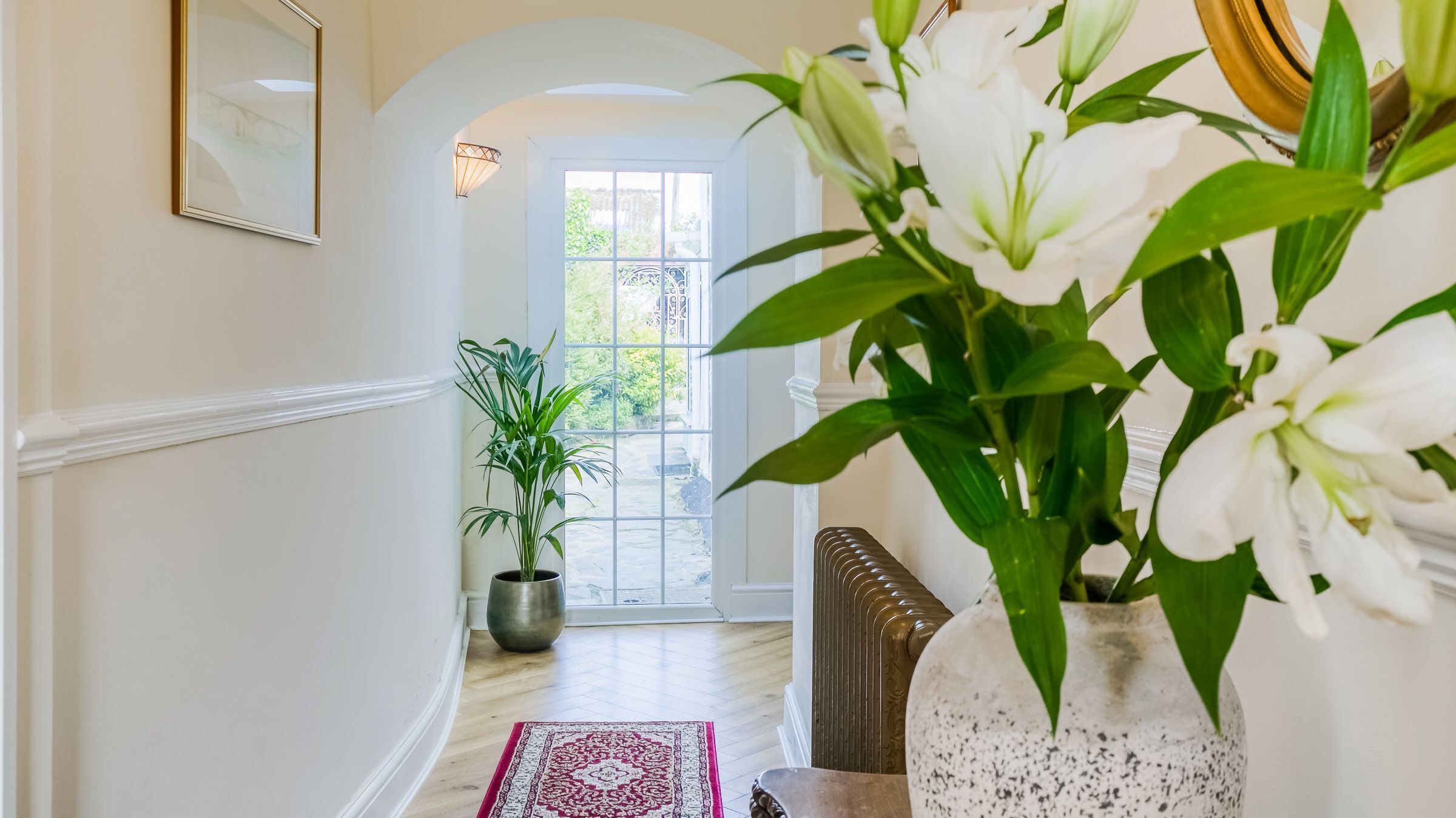 A hallway with a potted plant, a vase with flowers, and a door leading to a bright room.