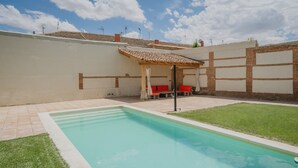 A rectangular pool with a tiled deck, a covered seating area with red cushions, and a grassy lawn.