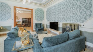A living room with a sofa, armchair, and TV, with patterned wallpaper and a radiator.