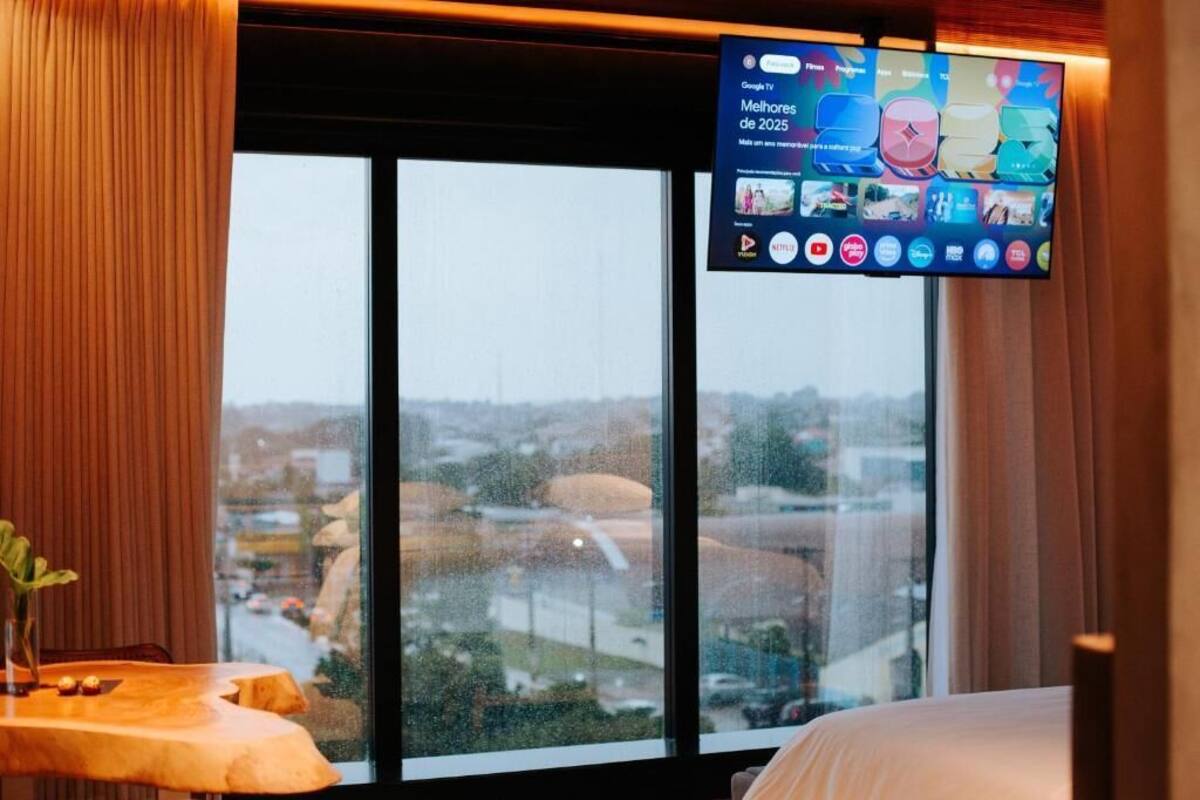 A hotel room with a large window, a bed, a bedside table with a vase, and a mounted television displaying various apps.
