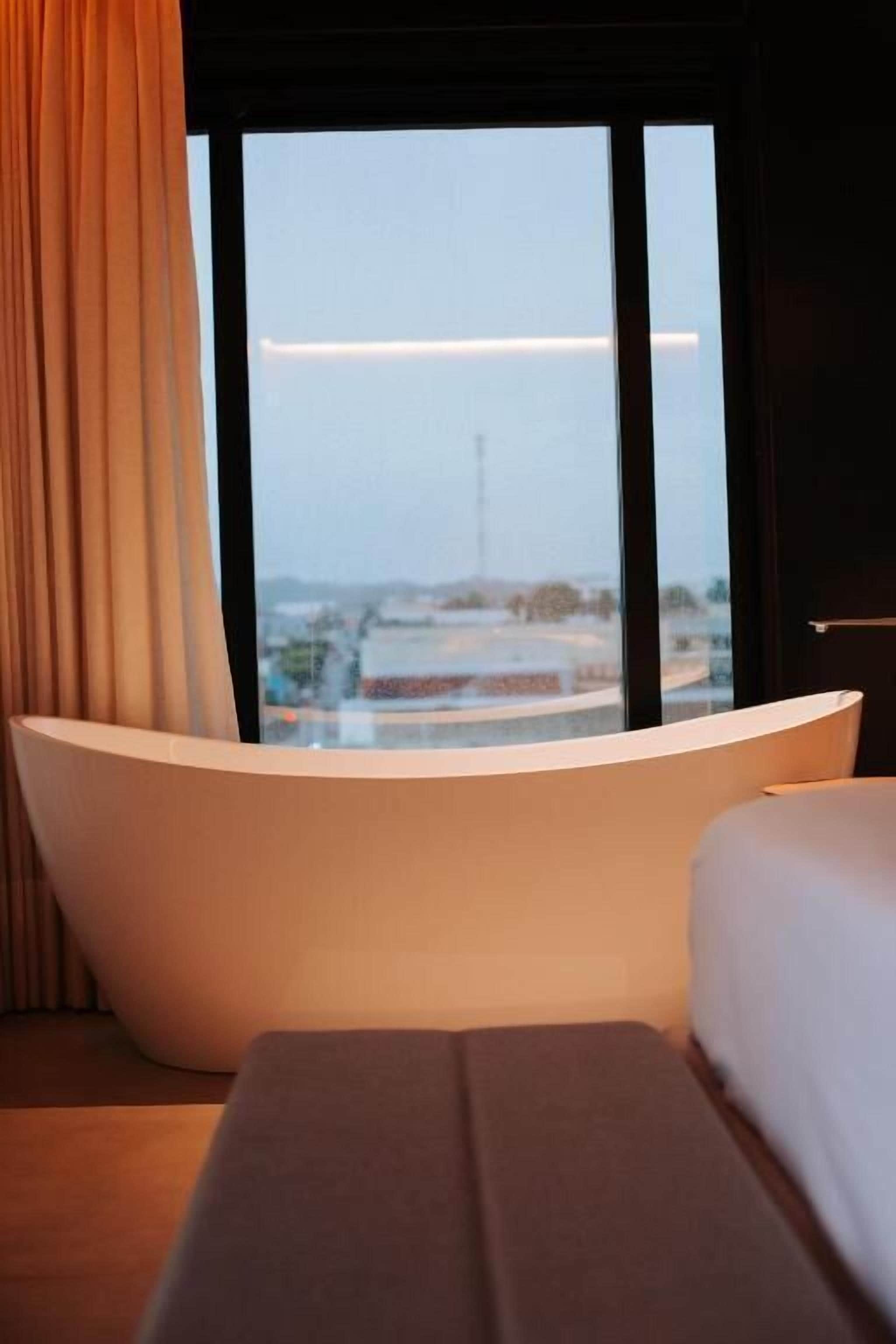 A modern hotel room with a large bathtub, a bed, and a view of a cityscape through a large window.
