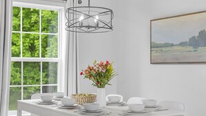 En matplats med ett vitt bord dukat för fyra personer, en vas med blommor, en geometrisk lampa och en landskapsmålning på väggen.