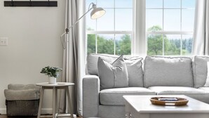 Ett vardagsrum med en soffa, ett litet bord, en golvlampa och ett fönster med utsikt över träd.