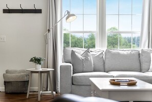 Un salon avec un canapé, une petite table, une lampe sur pied et une fenêtre donnant sur des arbres.