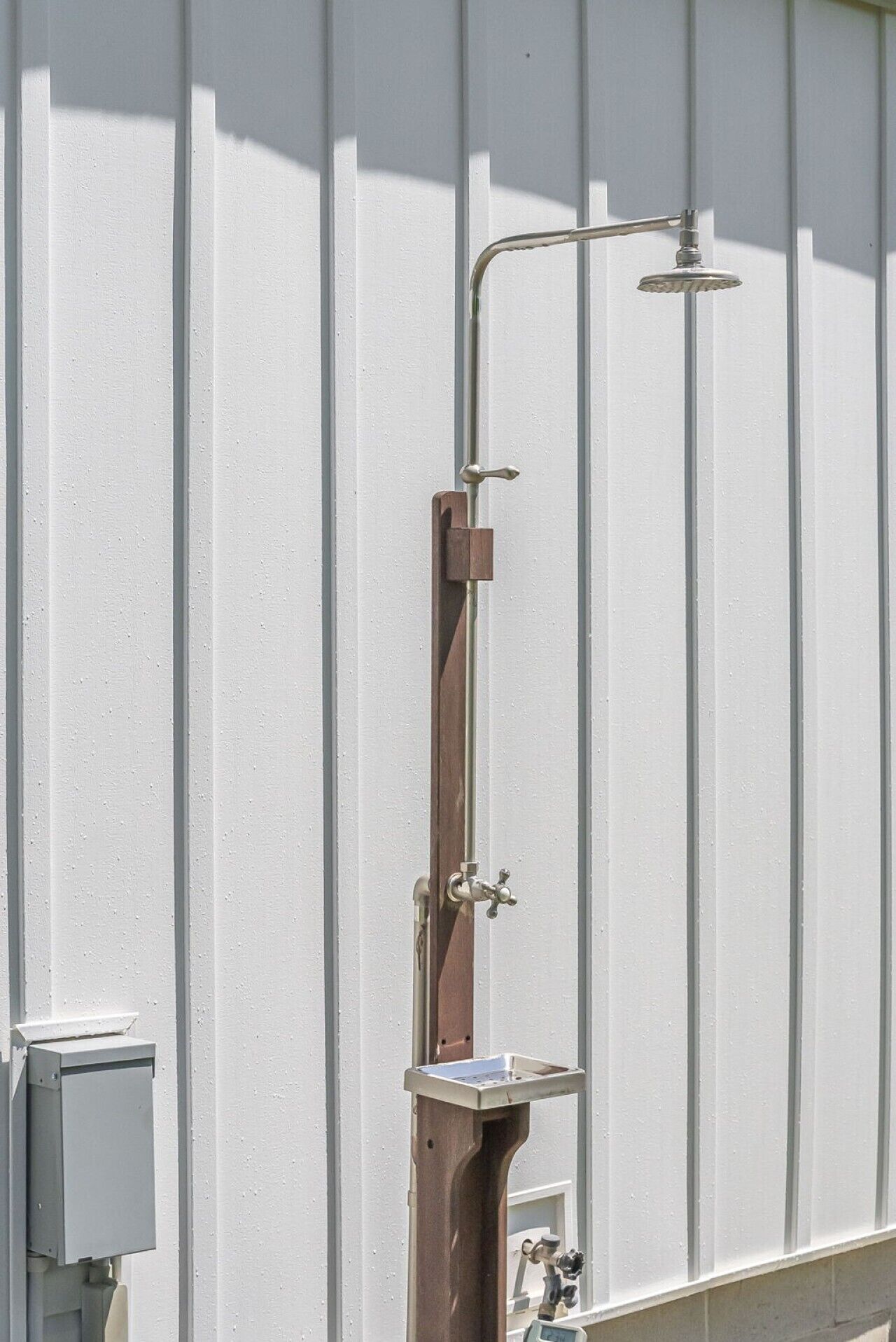 Un cabezal de ducha metálico unido a un tubo vertical, una estructura de soporte de madera y una caja metálica sobre una pared blanca.