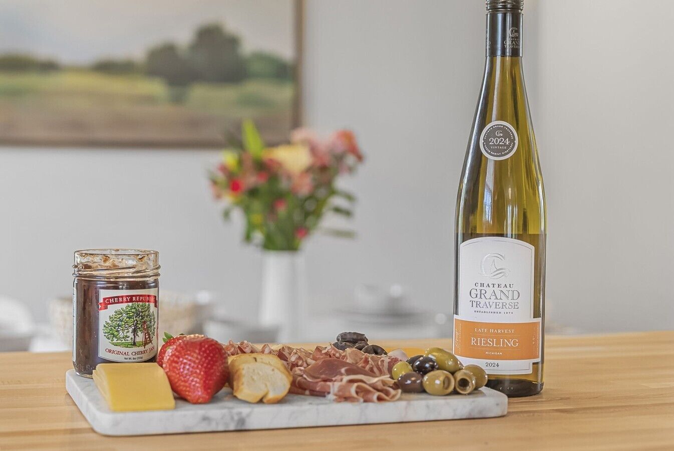 Una botella de vino Chateau Grand Traverse, un tarro de mermelada de cereza y una tabla de embutidos y quesos variados.