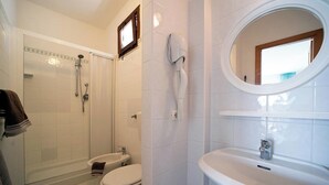 Een badkamer met douche, toilet en wastafel, met witte tegels en een raam boven de wastafel.