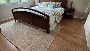 Una camera da letto con un letto in legno, un'ampia finestra, un comodino con una lampada e un tappeto sul pavimento.