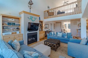 A spacious living area with a fireplace, a television, and a balcony with a railing. - Sun Realty pet-friendly oceanside Duck home w/ pool, hot tub & sound views (Duck)