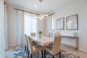 A dining room with a glass table, six chairs, a chandelier, and two abstract paintings on the wall. - Sleek Mid-Century Style Home in Rancho Mirage (Rancho Mirage)