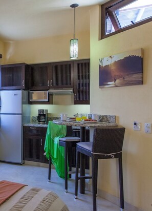 A compact kitchen with dark cabinets, a refrigerator, a microwave, and a dining area with two wicker chairs and a table. - Sayulita Cozy Studio, Fast Wi-Fi, Pool, Queen Bed, Kitchenette & Great Location (Sayulita)