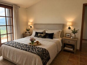 Una camera da letto con un letto grande, due comodini, un tavolino e una finestra con tende, con vista su piante.